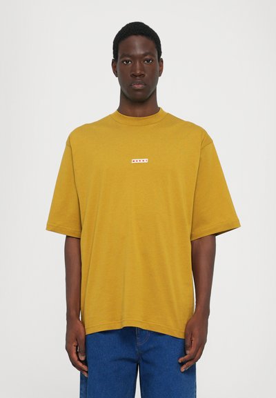 Senfgelbe T-Shirt mit kurzen Ärmeln, Rundhalsausschnitt und einem kleinen weißen Logo-Patch auf der Brust. Der Stoff wirkt glatt und leicht.