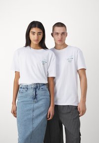 Camisetas de algodón blancas usadas por dos personas, con un pequeño diseño gráfico en el bolsillo. Una lleva una falda de mezclilla, la otra pantalones a rayas.