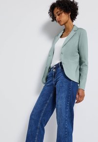 Blazer vert clair en tissu texturé, associé à un haut blanc et un jean large bleu. Doté de boutons lisses et d'un design ajusté.