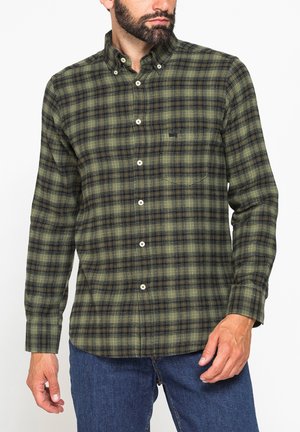 Uomo che indossa una camicia a quadri verde e nera con colletto abbottonato e jeans blu, in piedi contro uno sfondo semplice.