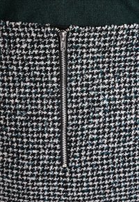 Vävt tyg i svart, vitt och teal med ett hundtandsmönster. Har en central silverfärgad dragkedja och en texturerad grön övre del.