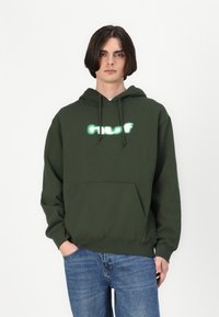 Hanorac verde din bumbac, cu un buzunar frontal, şnururi de strângere şi branding iluminat alb pe piept, purtat cu blugi blue.