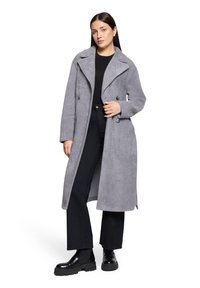 Manteau long en mélange de laine grise avec un col cranté, des boutons croisés et un tissu texturé. Porté sur un haut noir et un pantalon large.