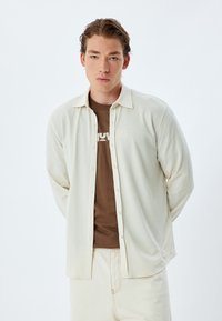 Camisa de manga larga de color beige claro, texturizada, con botones en la parte delantera y cuello, superpuesta a una camiseta marrón, llevada con pantalones de color crema.