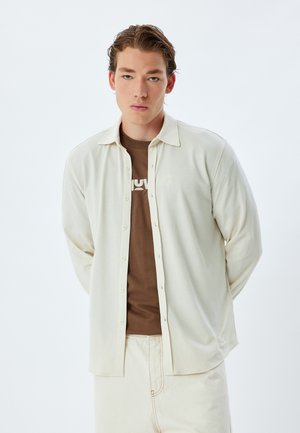 Camisa de manga larga de color beige claro, texturizada, con botones en la parte delantera y cuello, superpuesta a una camiseta marrón, llevada con pantalones de color crema.