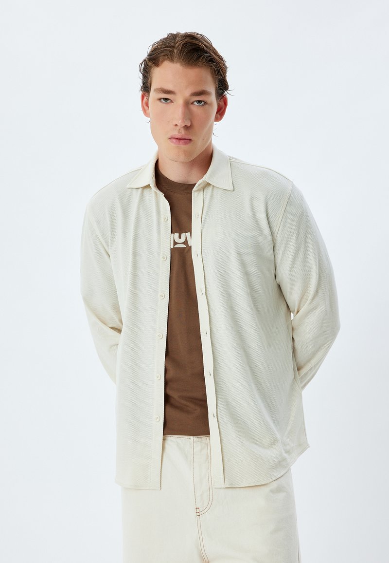 Vaalea beige teksturoitu pitkähihais shirt, jossa on napitettu etuosa ja kaulus, kerrostettuna ruskean t-paidan päälle, käytettynä kermaväristen housujen kanssa.
