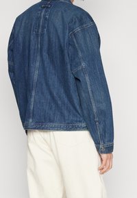 Denimjacka i mörkblått med avslappnad passform, stor baksömnad och knäppta ärmslut. Har synlig sömnad och minimalistisk design.