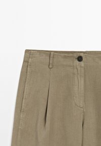 Pantalons khaki avec une texture lisse, des plis à l'avant et une fermeture à un seul bouton noir. Présente des passants de ceinture et des poches latérales.