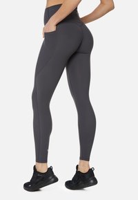 Grijze high-waisted leggings met een zijzak, gladde textuur en naaddetails. Gecombineerd met zwarte sportschoenen.