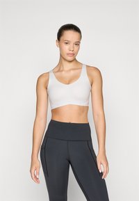 Siv modrček za šport z širokimi naramnicami in V-izrezom, izdelan iz gladkega blaga in z naramnicami pod prsmi. Usklajen z visokim črnim leggingom.