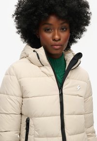 Pufferjacka i ljusbeige med blixtlås fram, svarta detaljer och huva. Har en quiltad textur och en liten vit logotyp på bröstet.