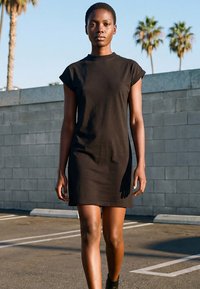 Frau, die im Freien geht, trägt ein schwarz kurzärmeliges Kleid und Stiefeletten; im Hintergrund sind Palmen und eine Betonwand zu sehen.