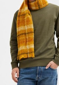 Vlněná šála v žluto-oranžovo-bílém károvaném vzoru, volně přehozená kolem krku, doplněná zeleným svetrem a modrými džínami.