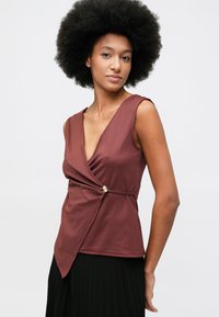 Γυναίκα με κούρεμα αφρού φορώντας αμάνικο τοπ σε ρουτζ απόχρωση με χρυσό κούμπωμα, συνδυασμένο με μαύρη πλισέ φούστα, σε λευκό φόντο.