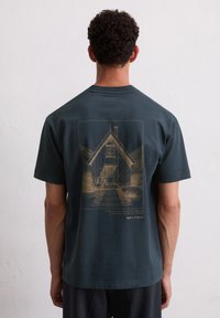 Dunkelblaues kurzärmliges T-Shirt mit einer goldfarbenen Illustration eines Hauses und Text, der für Umweltbewusstsein wirbt, sowie dem Markennamen.