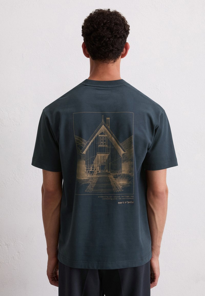Dunkelblaues kurzärmliges T-Shirt mit einer goldfarbenen Illustration eines Hauses und Text, der für Umweltbewusstsein wirbt, sowie dem Markennamen.