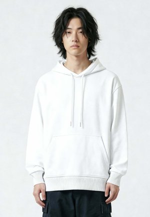 Giovane adulto con capelli neri di media lunghezza che indossa una felpa bianca semplice e pantaloni cargo neri, in piedi davanti a uno sfondo chiaro neutro.