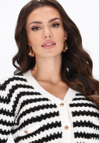 Cardigan cu dungi alb-negru, având guler în V, butoni aurii și țesătură texturată. Design clasic cu detalii ale buzunarului pe piept.