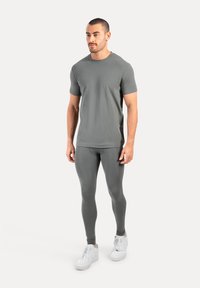 Man staat in een grijze T-shirt met korte mouwen, grijze leggings en witte sneakers tegen een effen lichte achtergrond.