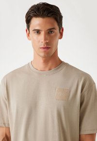 Beige bomulds T-shirt med rund halsudskæring, korte ærmer og et tekstureret lap med broderet logo på venstre brystområde.