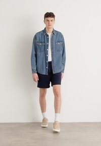 Camisa de denim en azul desgastado con dos bolsillos en el pecho, combinada con pantalones cortos marinos. El modelo lleva zapatillas beiges y calcetines blancos. Look sencillo y casual.