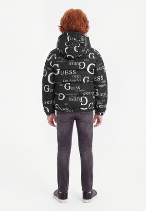 Enfant aux cheveux roux porte une doudoune noire avec le logo blanc GUESS imprimé, un jean foncé et des baskets noires, debout de dos sur fond blanc.