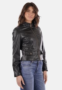 Giacca in pelle sintetica nera con colletto alto, zip frontale e due tasche anteriori. Silhouette aderente e texture liscia.