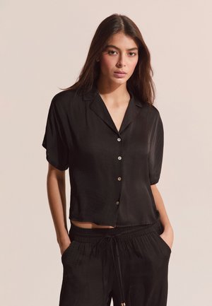 Jeune femme portant une chemise noire ample à manches courtes avec boutons et un pantalon assorti noir à cordon, debout les mains dans les poches.