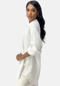 Chemise boutonnière blanche texturée à manches longues, avec une coupe décontractée et un ourlet arrondi, assortie à un pantalon de couleur claire.