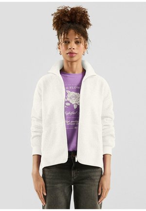 Frau mit lockigem Haar, die eine weiße Fleecejacke mit Reißverschluss über einem lila T-Shirt mit Blumenmuster und dunklen Jeans trägt und vor einem einfarbigen Hintergrund steht.