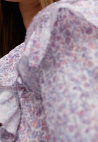 Leichte Bluse aus Stoff mit floralem Muster in Lila- und Rosatönen, mit Rüschendetails und einer zarten, leicht durchscheinenden Textur.