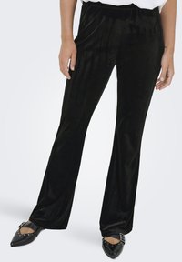 Pantaloni svasati in velluto nero con una texture liscia e una cintura elastica, abbinati a scarpe nere con punta e dettagli decorativi.