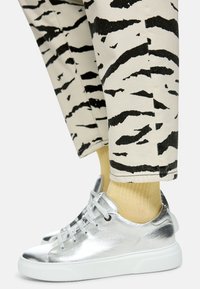 Silberne Metallic-Sneaker mit weißen Gummisohlen, kombiniert mit beigen Hosen mit schwarzem Tigerstreifenmuster und gelben, gerippten Socken.