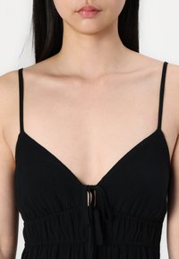 Vestido preto sem mangas com um decote profundo, tecido texturizado e um detalhe com cordão na cintura.