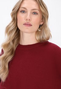 Burgunderfarbener Pullover mit rundem Ausschnitt und gerippter Textur, kombiniert mit goldenen Creolen, die doppelte Perlenakzente aufweisen.