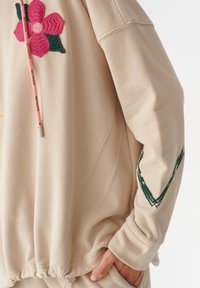 Sudadera beige con bordado floral en rosa y verde, cordones con logotipo y acento verde en la manga. Tejido suave y texturizado.