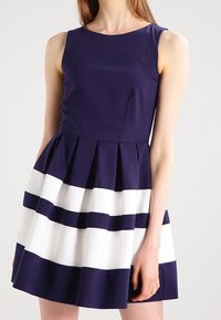 Robe sans manches bleu marine avec un corsage ajusté et une jupe évasée. Caractéristiques de rayures blanches horizontales et d'une texture plissée le long de la jupe.