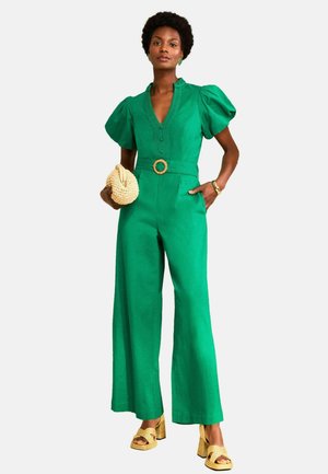 Frau trägt einen grünen Jumpsuit mit weitem Bein, Puffärmeln und Gürtel, hält eine beige gestrickte Clutch und trägt gelbe Sandalen mit Absatz.