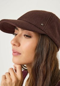 Casquette en feutre brun avec un pic recourbé, présentant des détails de coutures géométriques et un petit accent métallique sur le côté.