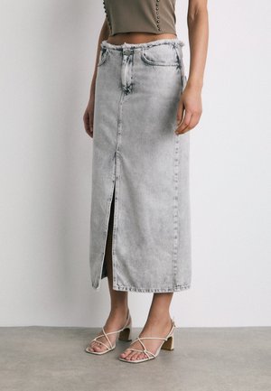 Personne portant une jupe midi en denim gris clair avec une fente à l'avant, associée à des sandales beiges à talons carrés et à brides, et un top court olive.