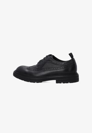 Zwarte leren brogue schoen met vleugelpunten perforaties, vetersluiting aan de voorkant, dikke zool en een trekbandje aan de hiel, weergegeven vanaf de zijkant op een witte achtergrond.