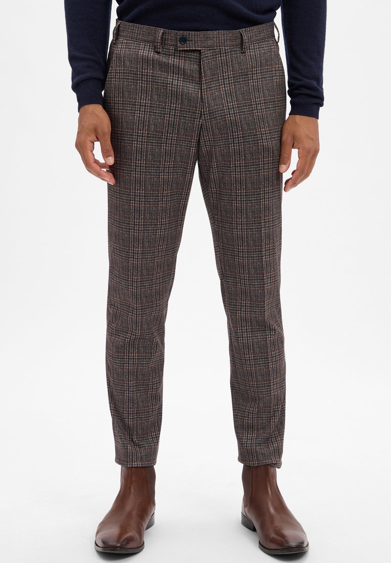 Checkered trousers in dark colors with a tailored fit, featuring subtle red and tan lines. Paired with brown leather boots.