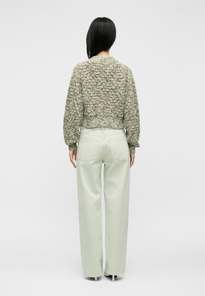 Femme aux cheveux noirs lisses portant un pull texturé vert et beige et un pantalon clair à jambes larges, debout de dos sur fond gris.