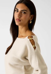 Beżowy sweter dziany z dekoltem w łódkę, z trzema złotymi akcentami na wycięciach na ramionach. Gładka tekstura, luźny krój.