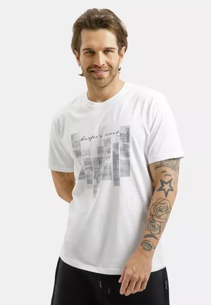 Uśmiechnięty mężczyzna z jasnobrązowymi włosami, ubrany w białą koszulkę z nadrukiem "Surfer's resort" i czarne spodnie, prezentujący tatuaże na lewym przedramieniu.