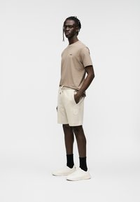 T-shirt beige avec logo, associé à un short beige clair avec une poche et un cordon de serrage, porté avec des chaussettes noires et des baskets blanches.