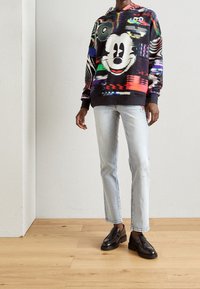 Sudadera con capucha colorida con patrones abstractos, que presenta un gran rostro de Mickey Mouse; combinada con vaqueros azul claro y mocasines negros.