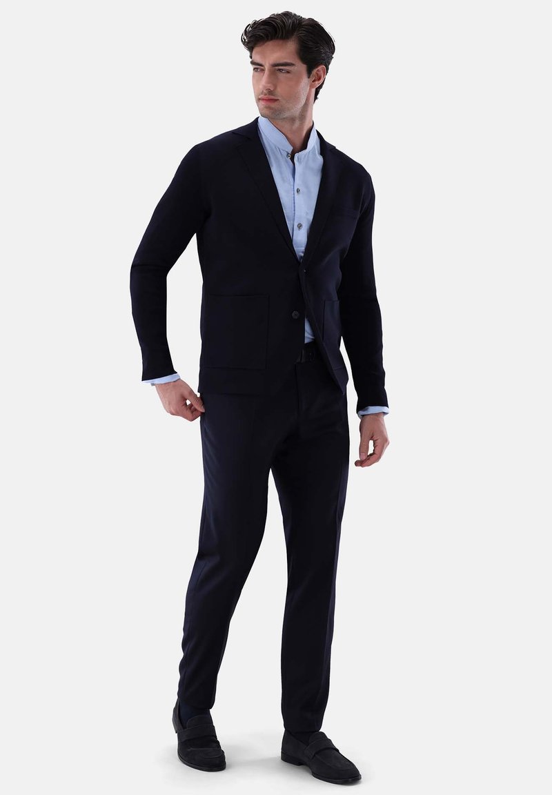 Marineblazer mit zwei vorderen Taschen, kombiniert mit einem hellblauen Hemd, dazu passende marinefarbene Hosen und schwarze Slipper. Glatte Textur.