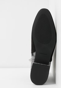 Semelle d'une chaussure plate à carreaux noirs et gris avec une semelle texturée et un talon carré, positionnée à la verticale sur un fond blanc.