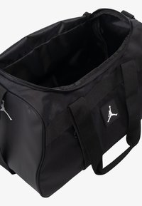 Black duffel bag made of durable fabric, featuring a smooth exterior, dual handles, and a logo patch on the side. Spacious open interior.